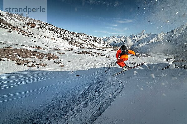 Ein männlicher Skisportler springt an einem sonnigen Tag von einem schneebedeckten Hang vor dem Hintergrund einer Berglandschaft mit schneebedeckten Bergen. Das Konzept des Wintersports