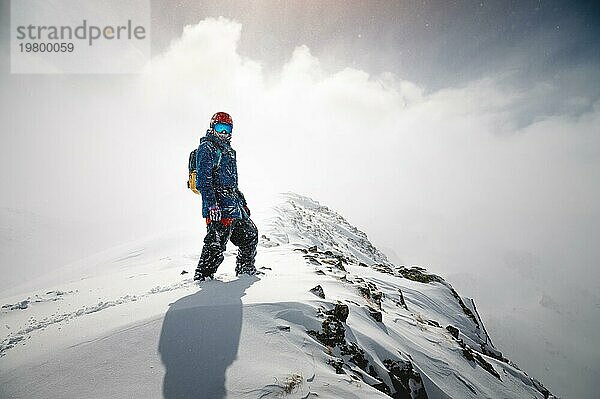 Porträt eines tapferen Mannes  der in Skikleidung auf einem verschneiten Berggipfel steht und mit seinem Rücken im Schneedunst die Sonne abschirmt