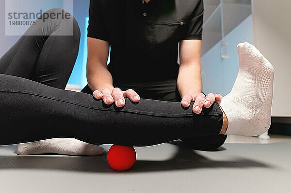 Myofasziale Entspannung. Ein männlicher Physiotherapeut setzt einen Ball ein  um die Beinmuskulatur eines Klienten im Sitzen mit einem roten Ball zu rehabilitieren. Das Konzept der Selbstmassage. Praktische Anwendung