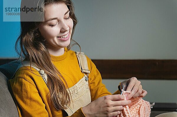 Junge glückliche kaukasische Frau sitzt lächelnd auf dem Sofa und häkelt Wollprodukte. Frauen Hobby Produktion von Kleidung aus Wolle