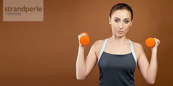 Panoramaaufnahme einer athletischen kaukasischen Frau mit Hanteln in ihren Händen. Steht auf einem braunen Hintergrund mit Hanteln in seinen Händen und schaut in die Kamera