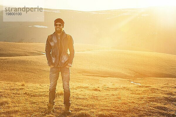 Modeporträt eines bärtigen jungen Mannes mit Sonnenbrille  Rucksack und Hut auf einem Hintergrund mit Copyspase in den Bergen bei Sonnenuntergang  Ein selbstbewusster Mann mit einem Bart