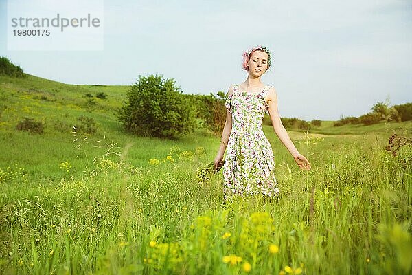 Junges glückliches lächelndes Mädchen in einem Kattunkleid mit einem Blumenstrauß geht eine Landstraße mit grünem Gras entlang