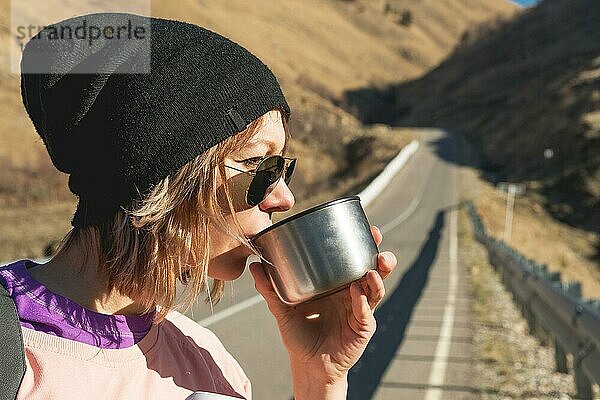 Ein Hipster Mädchen mit Sonnenbrille und schwarzem Hut trinkt Tee oder Kaffee aus einem Becher aus einer Thermoskanne in der Wildnis