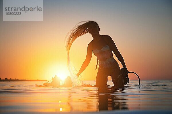 Silhouette Rahmen. Eine junge schlanke sexy weibliche Kitesurferin mit einem Brett und einem Kiteboard steht im Wasser im seichten Wasser bei Sonnenuntergang. Wassersport. Stilisierter Rahmen