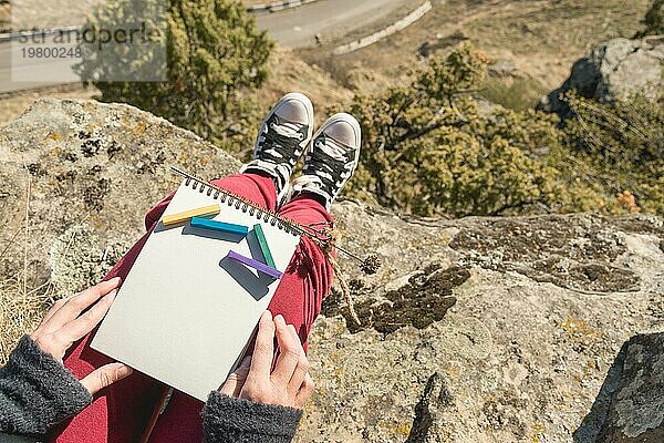 Blick aus der ersten Person auf den weiblichen Schoß ist ein Notizbuch mit Kreiden für die Zeichnung Pastelle. Weibliche Hände halten ein Notizbuch in der Natur. Ein Mädchen  das auf einer Klippe in der Nähe einer Klippe sitzt  malt eine Pastelllandschaft. Beschaffung für den Designer