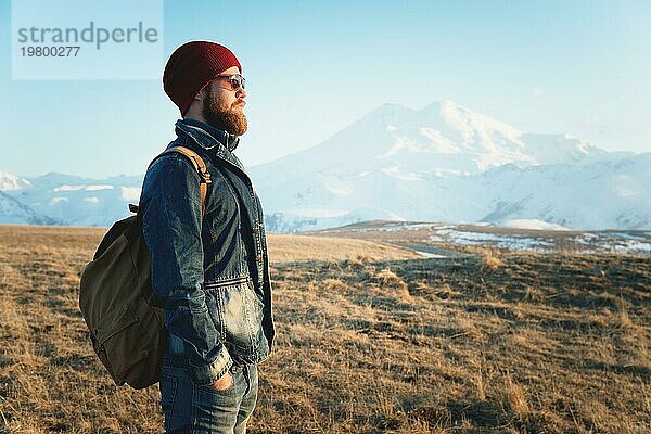 Porträt eines nachdenklichen stilvollen Hipsters mit Bart  Sonnenbrille und Hut mit einem Rucksack vor dem Hintergrund des schneebedeckten Berges Elbrus im Kaukasus bei Sonnenuntergang