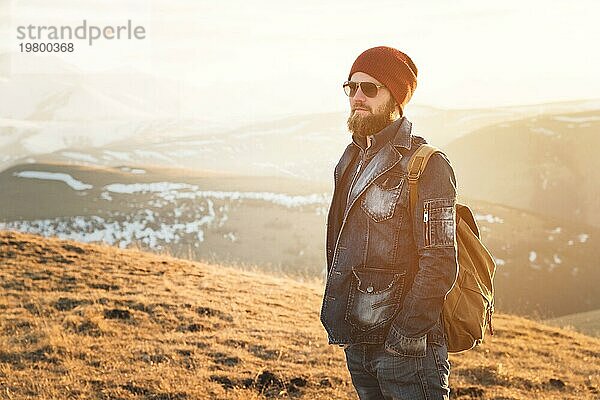 Porträt eines nachdenklichen stilvollen Hipsters mit Bart  Sonnenbrille und Hut mit einem Rucksack vor dem Hintergrund des schneebedeckten Berges Elbrus im Kaukasus bei Sonnenuntergang