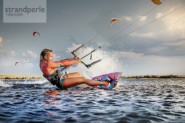 Professionelle Kitesurfer junge kaukasische Frau gleitet auf einem Brett entlang der Meeresoberfläche bei Sonnenuntergang vor dem Hintergrund der schönen Wolken und andere Drachen. Aktiver Wassersport