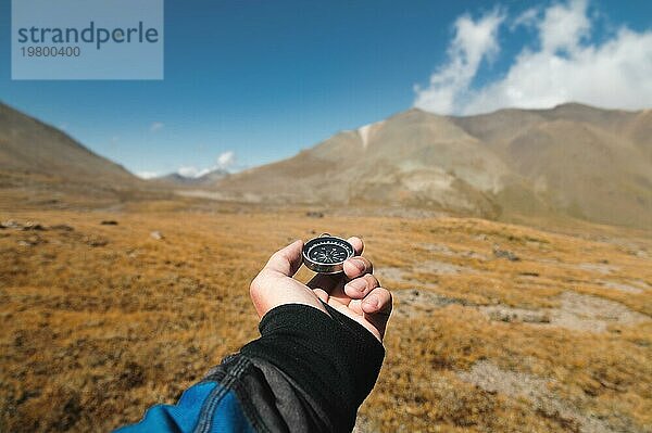 Weitwinkel POV Aufnahme eines männlichen Reisenden  der einen Magnetkompass vor dem Hintergrund einer bergigen Landschaft in der Hand hält. Orientierung und Finden des Weges