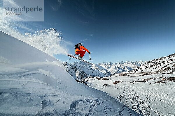 Ein männlicher Skisportler springt an einem sonnigen Tag von einem schneebedeckten Hang vor dem Hintergrund einer Berglandschaft mit schneebedeckten Bergen. Das Konzept des Wintersports