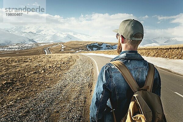 Ein bärtiger Mann mit einer Mütze und einem Rucksack  der bereit ist  einen langen Weg zu gehen. Ein Mann auf einer Landstraße vor der Kulisse von Bergen und Wolken