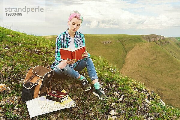 Ein reisendes Mädchen sitzt in den Bergen im Gras und liest ein Buch vor dem Hintergrund epischer Berge. Das Konzept des Lesens während der Erholung und des Urlaubs