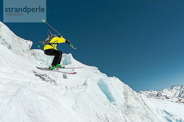 Ein Skifahrer in voller Sportausrüstung springt von der Spitze des Gletschers in den Abgrund  vor dem Hintergrund des blaün Himmels und der schneebedeckten Berge des Kaukasus. Blick von hinten. Elbrus Gebiet. Russland