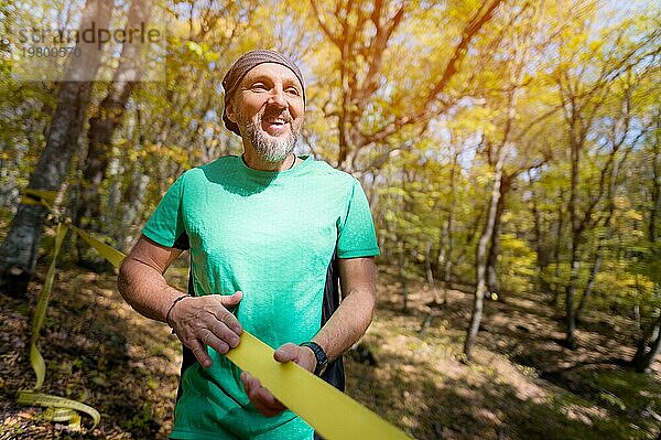 Porträt eines reifen Sportlers mit Bart neben einer gespannten Slackline im herbstlichen Wald am Nachmittag. Das Konzept des Sports nach 40 Jahren