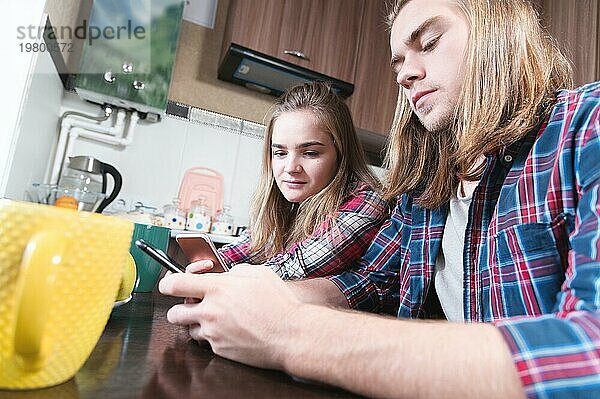 Ein junges  lächelndes Paar  ein Mann und ein Mädchen mit langen Haaren  sitzen am Esstisch in der Küche und benutzen Telefone  um in Chatrooms und sozialen Netzwerken zu kommunizieren. Probleme der Internetabhängigkeit in jungen Familien