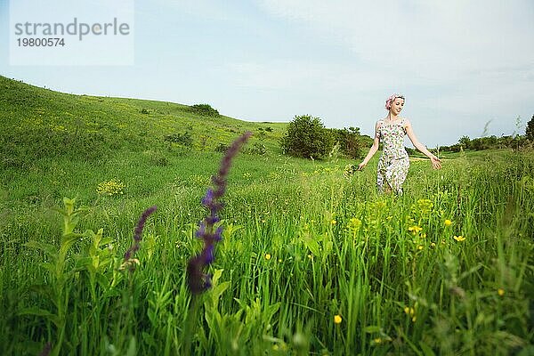Junges glückliches lächelndes Mädchen in einem Kattunkleid mit einem Blumenstrauß geht eine Landstraße mit grünem Gras entlang