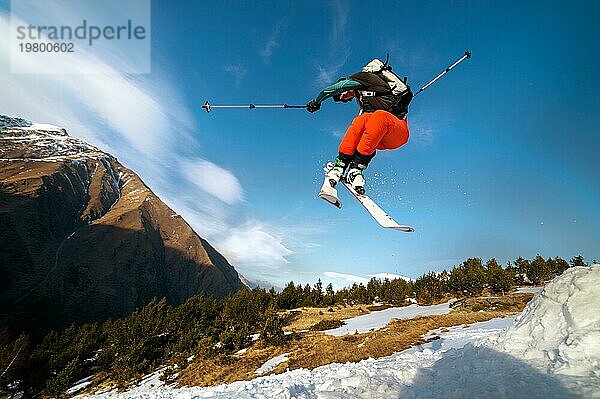 Mann Skifahrer im Flug nach dem Sprung von einem Kicker im Frühjahr vor dem Hintergrund der Berge und blaün Himmel. Nahaufnahme mit Weitwinkel. Das Konzept der Schließung der Skisaison und Skifahren im Frühjahr