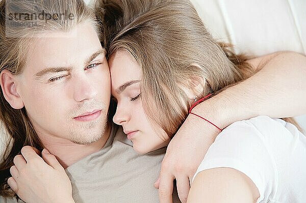 Blick von oben auf einen jungen Mann und eine Frau  die in einem weißen Bett liegen und sich umarmen  wobei der Mann verstohlen in die Kamera schaut  während die Frau schläft. Das Konzept der Geheimnisse im Familienleben und gesunde sexuelle Beziehungen