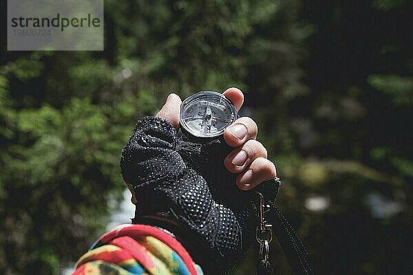Eine behandschuhte Hand hält einen Magnetkompass vor dem Hintergrund eines Nadelwaldes. Das Konzept der Outdoor Navigation