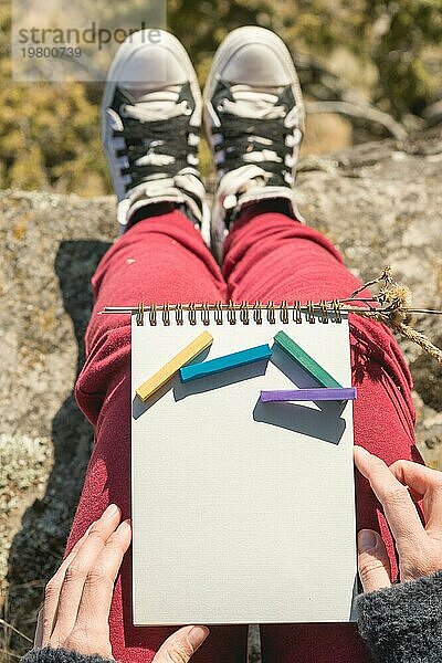 Blick aus der ersten Person auf den weiblichen Schoß ist ein Notizbuch mit Kreiden für die Zeichnung Pastelle. Weibliche Hände halten ein Notizbuch in der Natur. Ein Mädchen  das auf einer Klippe in der Nähe einer Klippe sitzt  malt eine Pastelllandschaft. Beschaffung für den Designer