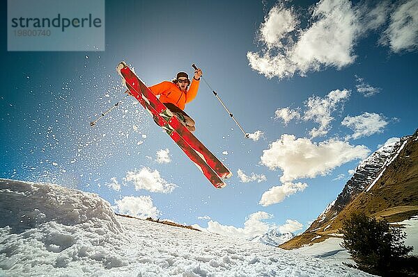 Schöne junge kaukasische Mann in einer orangefarbenen Jacke und schwarzen Shorts springen von einem Sprungbrett auf roten Skiern vor dem Hintergrund der Berge  Himmel  Wolken  Sonne