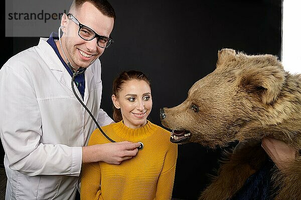 Lächelnder junger Arzt mit Stethoskop und Patient  der einen Bären untersucht  Studio für medizinischen Humor  der entscheidet  wen er behandelt  Mensch oder Tier