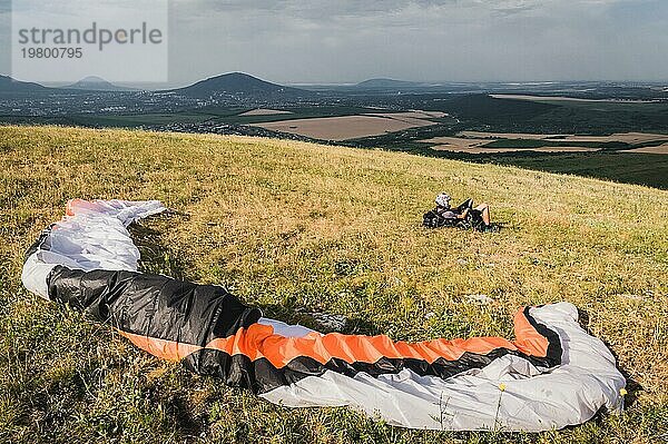 Ein professioneller Gleitschirmflieger in voller Ausrüstung und mit Helm liegt auf dem Gras hoch in den Bergen und schaut in die Wolken