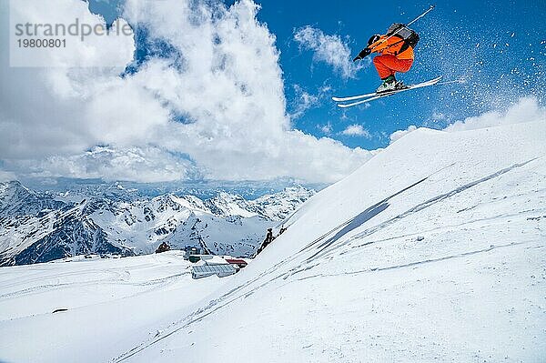 Ein männlicher Skifahrer in einem orangefarbenen Anzug fliegt durch die Luft  nachdem er an einem sonnigen Tag von einem Schneefeger hoch in den kaukasischen Bergen gesprungen ist  inmitten schneebedeckter Berge mit blauem Himmel und weißen Wolken