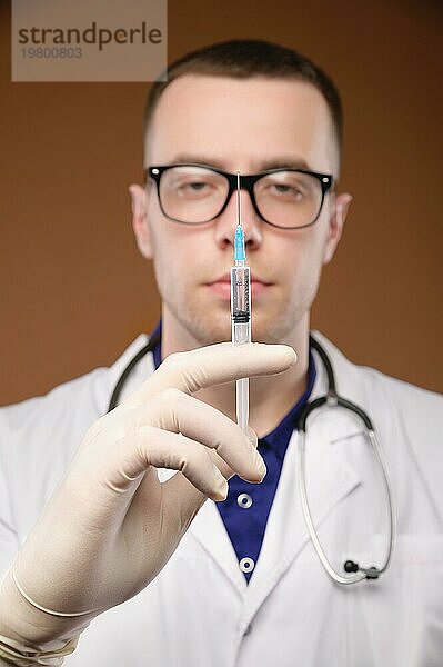 Ein junger männlicher Arzt mit Brille  Stethoskop um den Hals und weißem Kittel hält eine Plastikspritze mit einer Injektion in der Hand. Fokus auf die Spritze