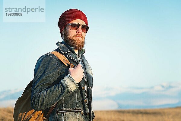 Porträt eines nachdenklichen stilvollen Hipsters mit Bart  Sonnenbrille und Hut mit einem Rucksack vor dem Hintergrund des schneebedeckten Berges Elbrus im Kaukasus bei Sonnenuntergang