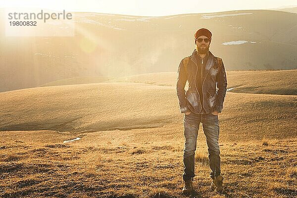 Modeporträt eines bärtigen jungen Mannes mit Sonnenbrille  Rucksack und Hut auf einem Hintergrund mit Copyspase in den Bergen bei Sonnenuntergang  Ein selbstbewusster Mann mit einem Bart