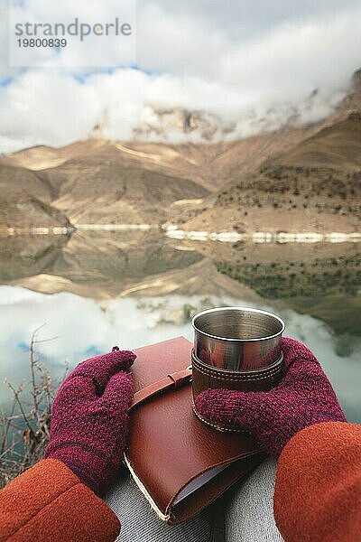 Eine Ichperspektive von Frauenhänden in Mänteln und Fäustlingen  die einen Metallbecher mit Tee oder Kaffee und ein ledernes Taschenkalenderbuch vor dem Hintergrund eines Bergsees und Felsen in den Wolken halten. Reisekonzept