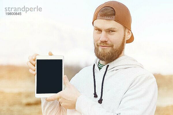 Nahaufnahme eines lächelnden Hipsters in einer braunen Mütze  der im Freien einen weißen Tabletcomputer mit einem ausgeschnittenen Feld für den Designer hält. Ein bärtiger Mann schaut auf das Tablet. Hintergrund unscharf