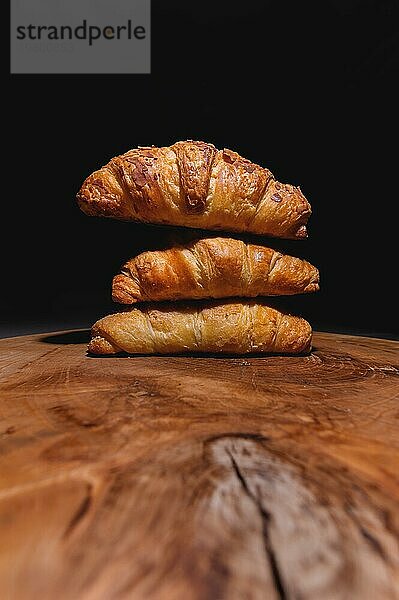 Nahaufnahme eines Stapels von drei Croissants auf einem Holzbrett vor einem dunklen Hintergrund. Leckeres und gesundes Frühstück