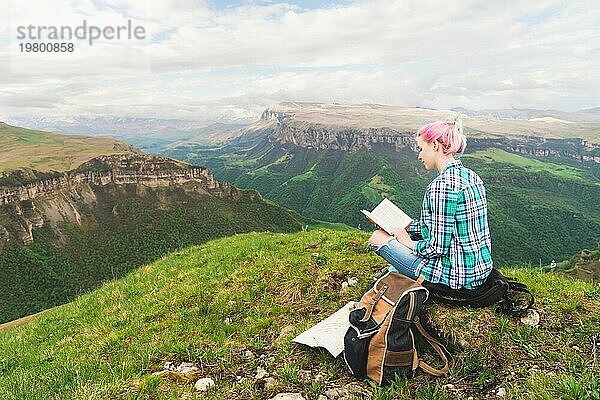 Ein reisendes Mädchen sitzt in den Bergen im Gras und liest ein Buch vor dem Hintergrund epischer Berge. Das Konzept des Lesens während der Erholung und des Urlaubs