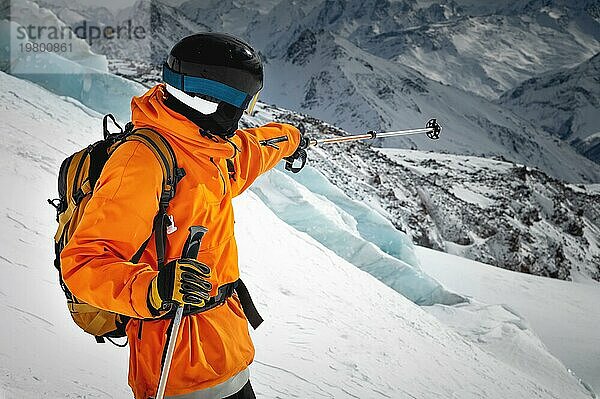 Ein professioneller Skifahrer in roter Jacke  Helm und Maske steht auf einer Piste vor der Kulisse eines Gletschers und epischer Berge und zeigt mit einem Skistock auf eine Bergkette Freeride Konzept