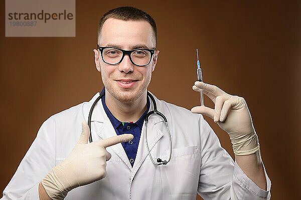 Ein junger kaukasischer Arzt im weißen Kittel und mit Brille hält eine Spritze mit einem Impfstoff. Ein freundlicher  knuddeliger junger Mann zeigt mit dem Finger auf eine Spritze und bietet an  sich impfen zu lassen