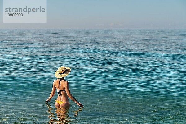 Rückenansicht einer schlanken kaukasischen jungen Frau  die in einem Badeanzug und einem Strohhut hüfttief im ruhigen transparenten Meerwasser steht