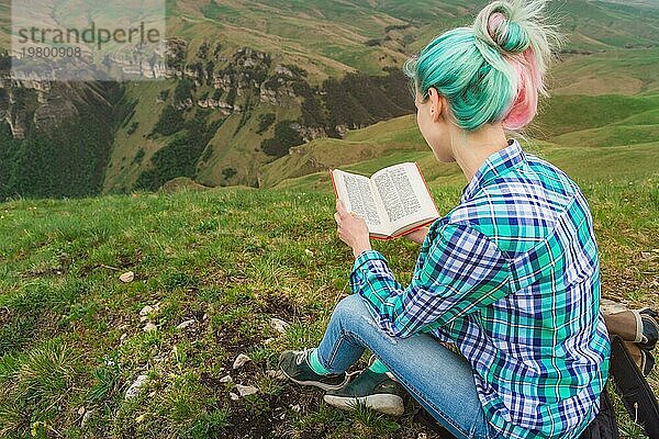 Ein reisendes Mädchen sitzt in den Bergen im Gras und liest ein Buch vor dem Hintergrund epischer Berge. Das Konzept des Lesens während der Erholung und des Urlaubs