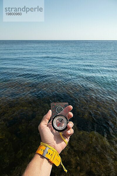 Die Hand eines Mannes mit Armbanduhr hält einen Magnetkompass vor dem Hintergrund des Meeres