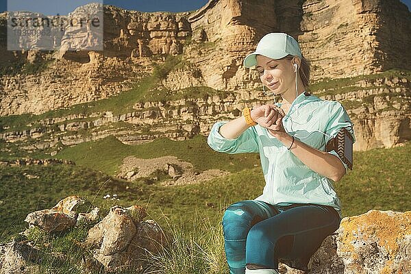 Porträt eines lächelnden jungen Fitness Mädchens mit Mütze und Kopfhörern  das auf einem Felsen sitzend im Freien vor einem Felsenhintergrund ihre intelligente Uhr überprüft