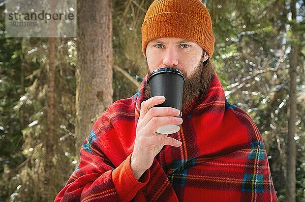 Bärtiger kaukasischer Mann mit Hut  eingewickelt in eine Decke  mit einem Becher mit heißem Getränk in einem Pappbecher. in einem winterlichen Nadelwald an einem sonnigen Tag