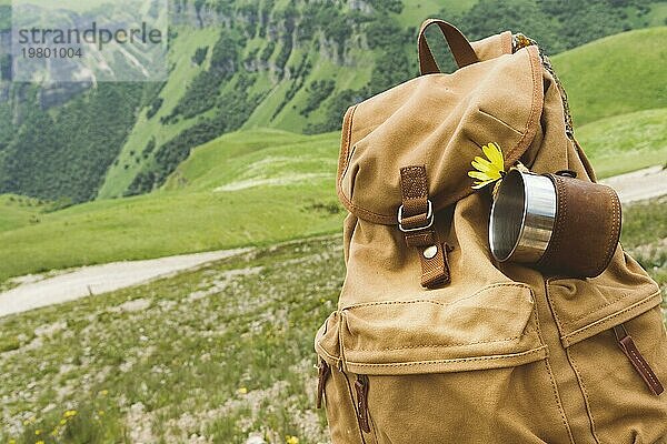 Hipster gelben Vintage Rucksack mit einem Becher auf sie mit einem Becher Nahaufnahme Vorderansicht befestigt. Traveler's Reisetasche im Hintergrund einer Berglandschaft