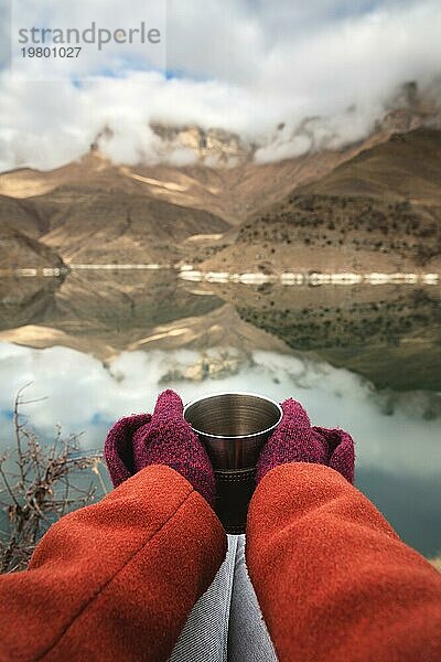 Eine Ichperspektive von Frauenhänden in Mänteln und Fäustlingen  die einen Metallbecher mit Tee oder Kaffee vor dem Hintergrund eines Bergsees und Felsen in den Wolken halten. Reisekonzept