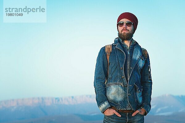 Modeporträt eines bärtigen jungen Mannes mit Sonnenbrille  Rucksack und Hut auf einem Hintergrund mit Copyspase in den Bergen bei Sonnenuntergang  Ein selbstbewusster Mann mit einem Bart