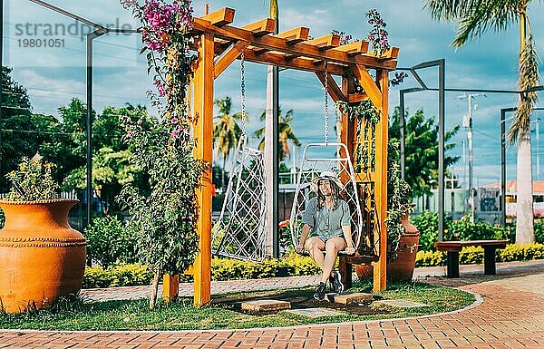 Schönes Mädchen mit Hut sitzt auf einer weißen Schaukel in einem schönen Garten. Porträt eines lächelnden Mädchens  das auf einer Schaukel in La Calzada  Granada  Nicaragua sitzt