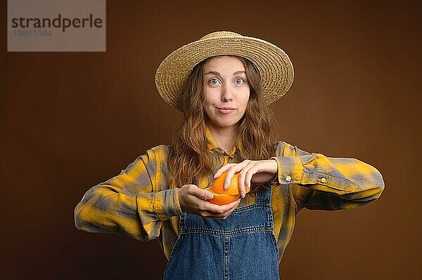 Attraktive rustikale kaukasische junge Frau mit Strohhut  die geschnittene Orangenfrüchte hält. Studioporträt vor braunem Hintergrund. Schaut in die Kamera