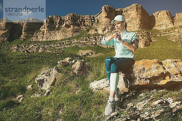 Porträt eines lächelnden jungen Fitness Mädchens mit Mütze und Kopfhörern  das auf einem Felsen sitzend im Freien vor einem Felsenhintergrund ihre intelligente Uhr überprüft