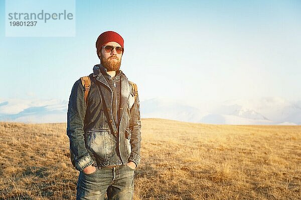 Porträt eines nachdenklichen stilvollen Hipsters mit Bart  Sonnenbrille und Hut mit einem Rucksack vor dem Hintergrund des schneebedeckten Berges Elbrus im Kaukasus bei Sonnenuntergang